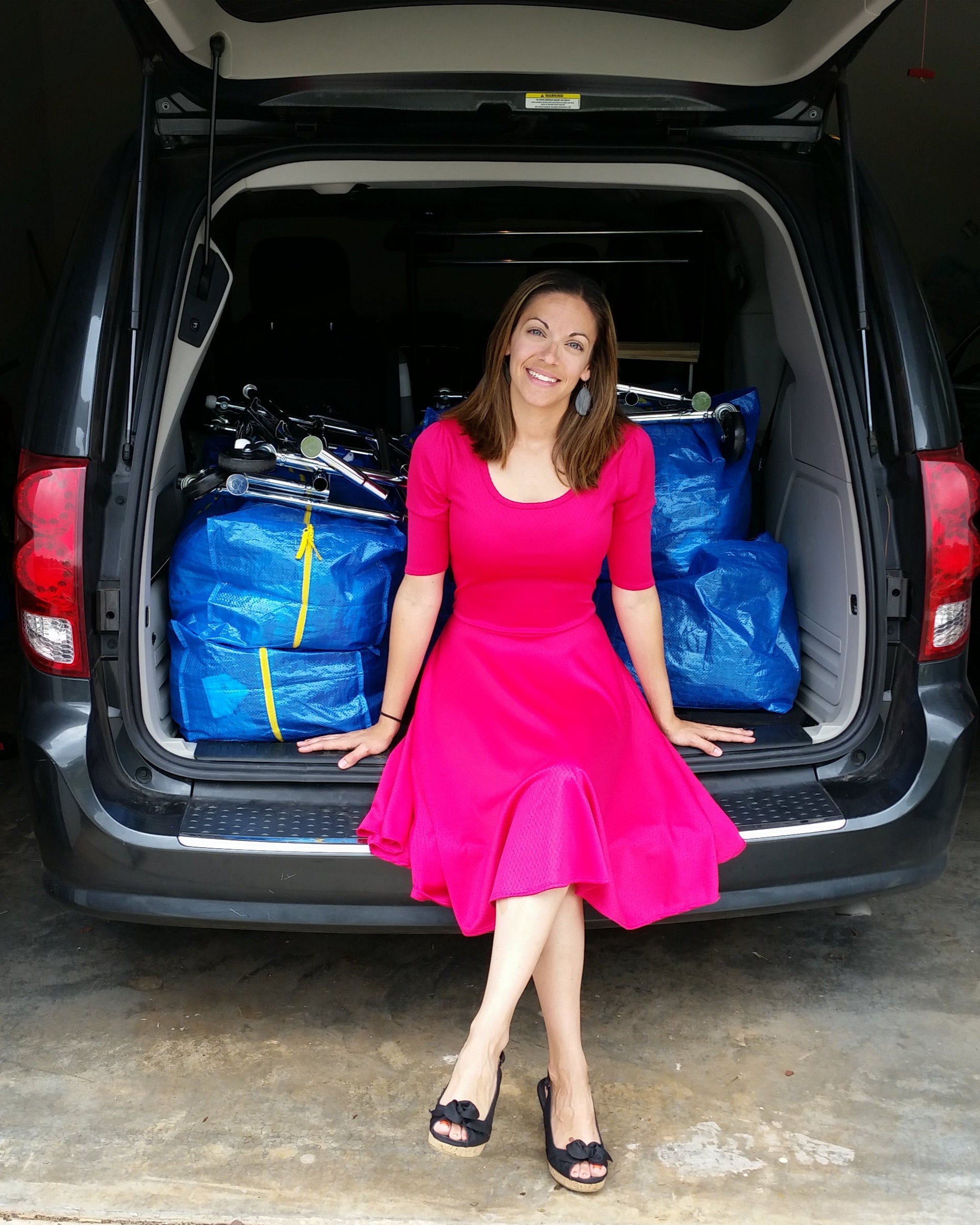 Woman in a pink dress sitting on the tailgate of a van filled with blue bags.